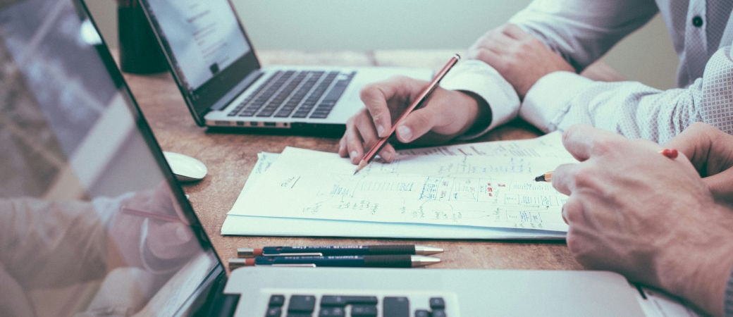 Business professionals collaborating at a desk with laptop and paper documents, hands visible writing notes during meeting