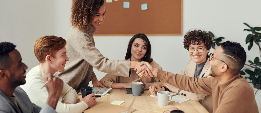 Diverse group of young professionals in meeting room, with two people shaking hands across table while others smile and observe