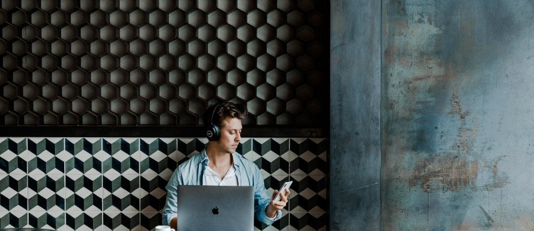 Person wearing headphones working on MacBook laptop while checking smartphone, seated against geometric patterned wall in modern space
