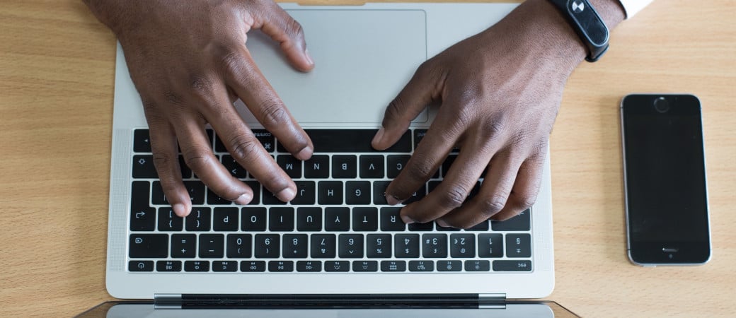 Person typing on laptop keyboard with smartphone nearby on wooden desk, depicting digital access to reading materials