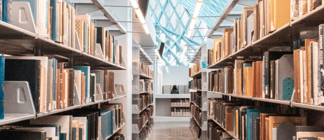Modern library interior with rows of bookshelves filled with physical books, featuring a distinctive glass ceiling allowing natural light to illuminate the space